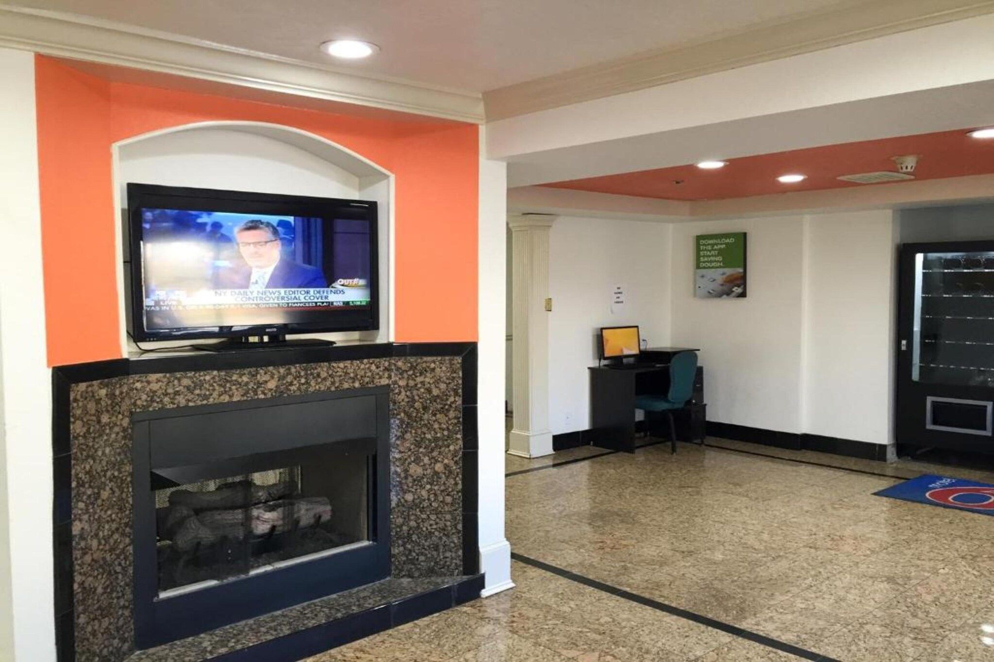 A lobby with a television displaying news, a fireplace, a desk with a computer, and a door with a poster.