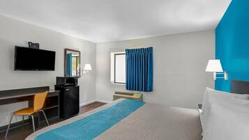 A hotel room with a bed, a desk with a chair, a television, a microwave, a window with curtains, and a wall-mounted lamp.
