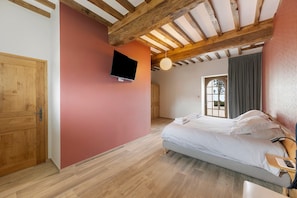 Ein Schlafzimmer mit Bett, einem an der Wand befestigten Fernseher, Holzbalken an der Decke und einem Fenster mit Vorhängen.