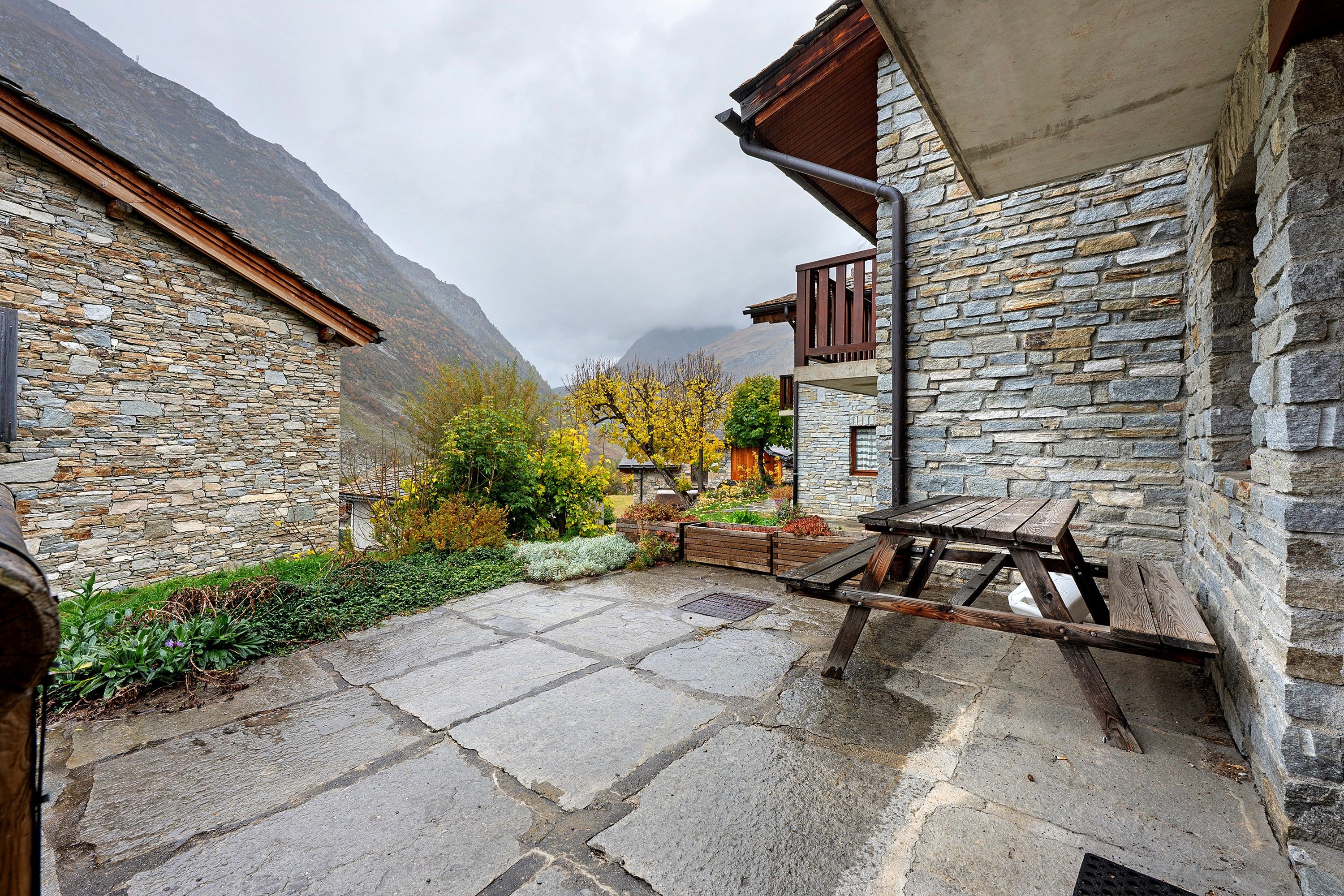 A stone building with a wooden balcony, a stone wall, and a paved courtyard with a wooden bench.