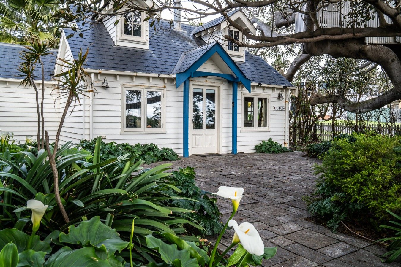 A white house with a blue door and windows, surrounded by lush greenery and a paved walkway.