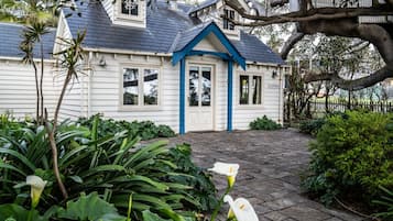 A white house with a blue door and windows, surrounded by lush greenery and a paved walkway.