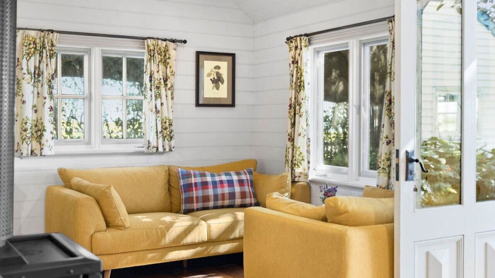A room with a yellow sofa, floral curtains, and a framed botanical print on the wall.