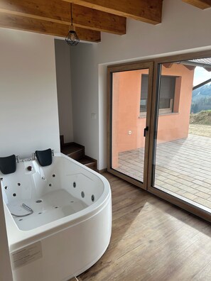 A modern bathroom with a large white bathtub, wooden flooring, and a glass door leading to an outdoor deck. - Cozy 2-bedroom house in charming Gradišča with AC and Sauna (Cirkulane)