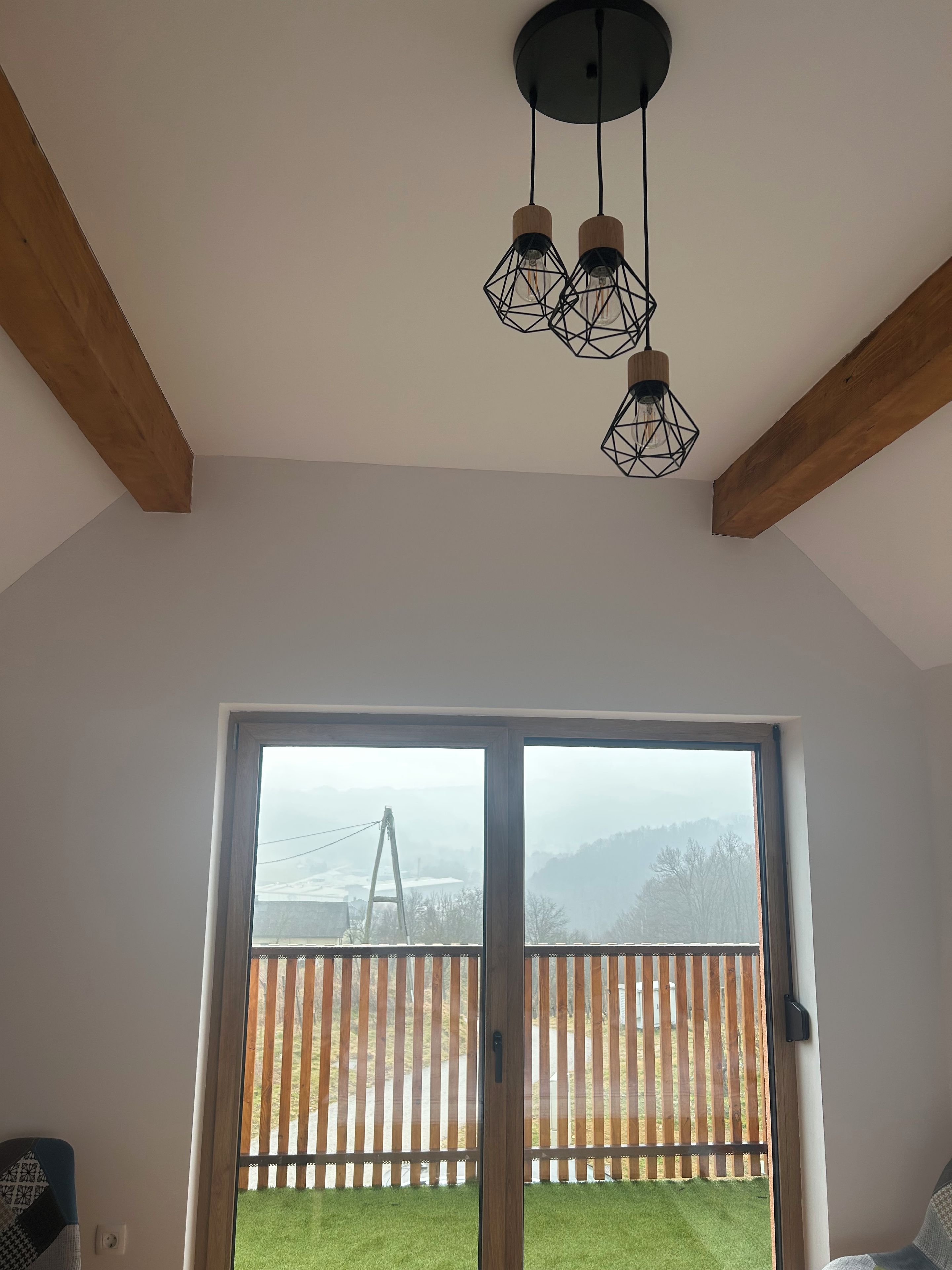 A room with a wooden ceiling beam, a sliding glass door leading to a balcony with a view of a wooden fence and distant hills.