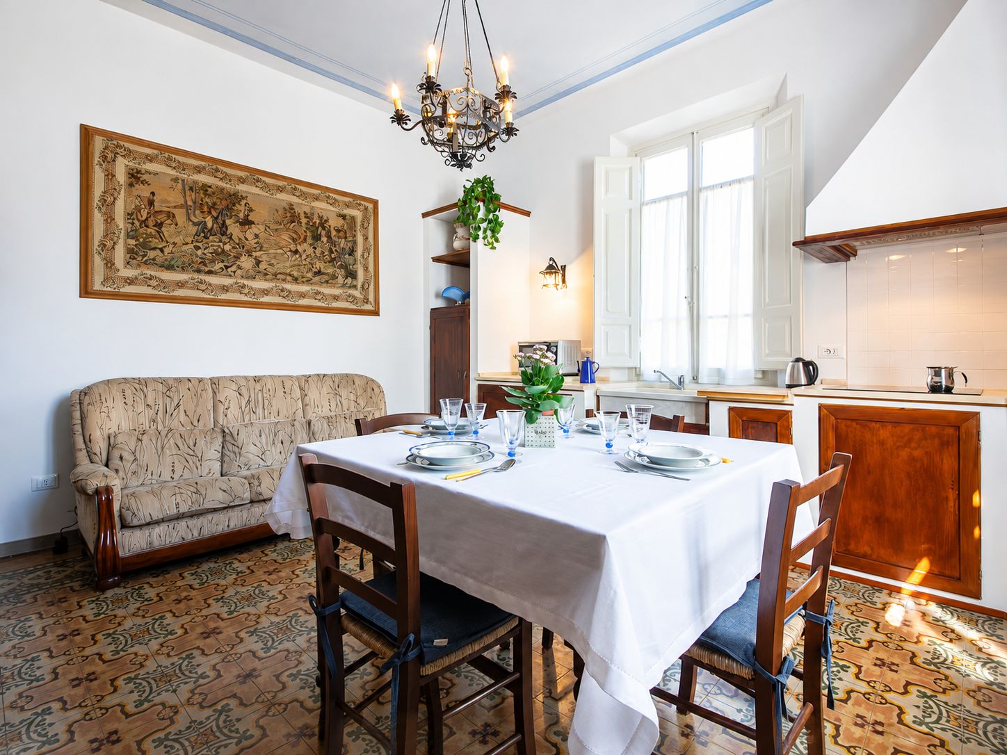 A dining room with a table set for four, a sofa, a chandelier, and a large painting on the wall.