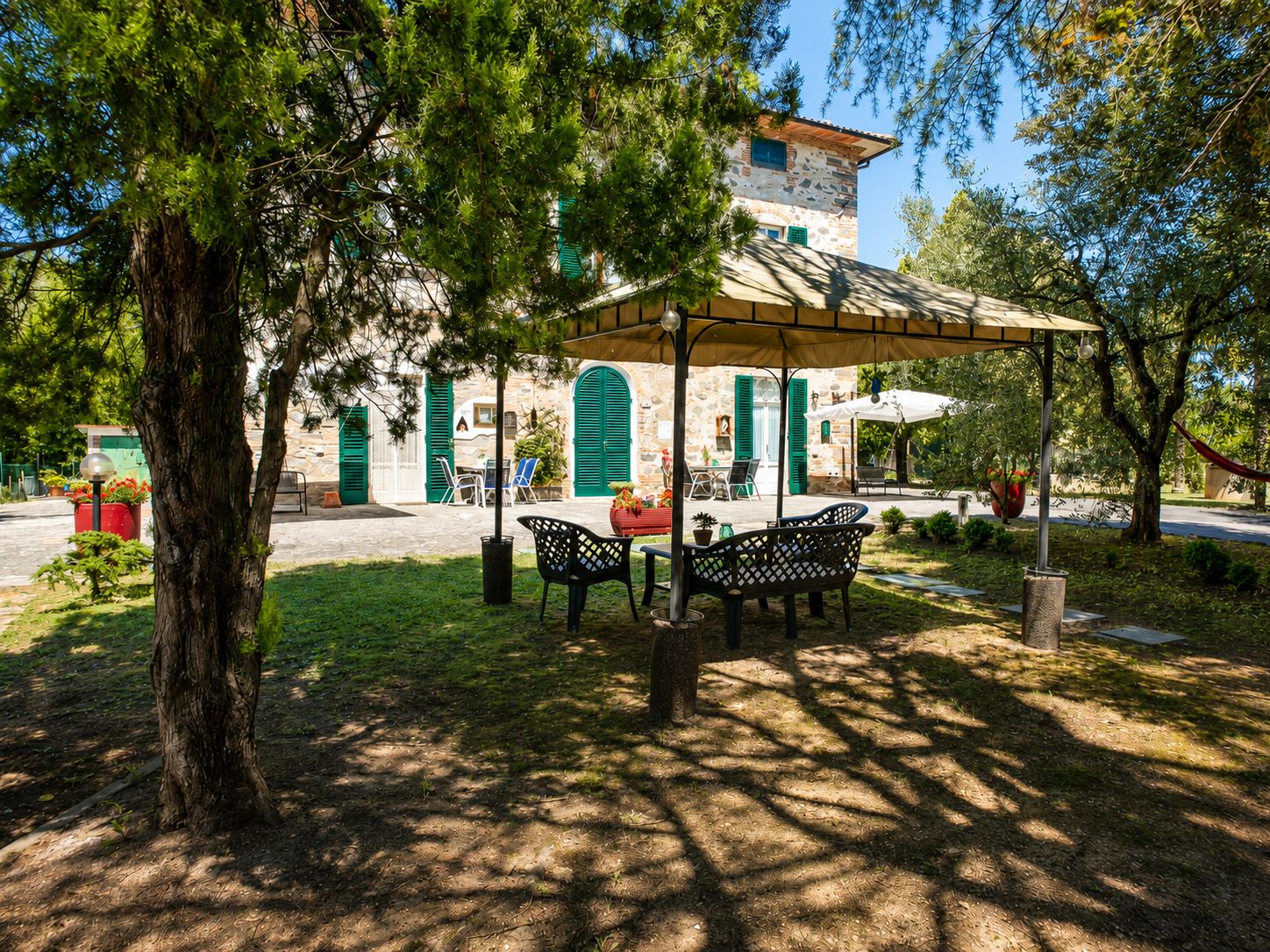 Un patio al aire libre con un edificio de piedra al fondo, una zona de estar cubierta y un árbol en primer plano.