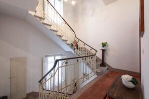 Una escalera con barandilla de metal que sube a un piso superior, una planta en maceta sobre una mesa de madera y una puerta a la izquierda.