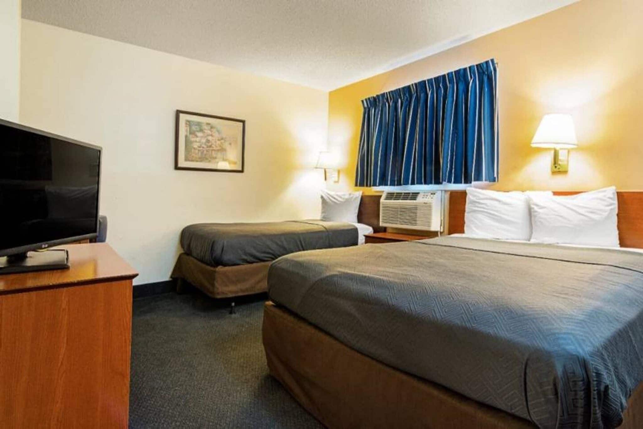 A hotel room with a bed, a desk with a television, a bedside table, and a window with curtains.