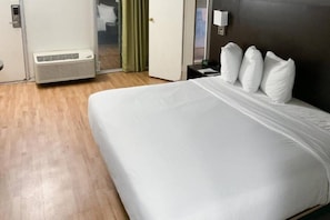 A hotel room with a large bed, a bedside table with a lamp, and a wall-mounted air conditioning unit. - 1 King Bed | Mobil Access Tub Nsmk Kitchen (Columbus GA)