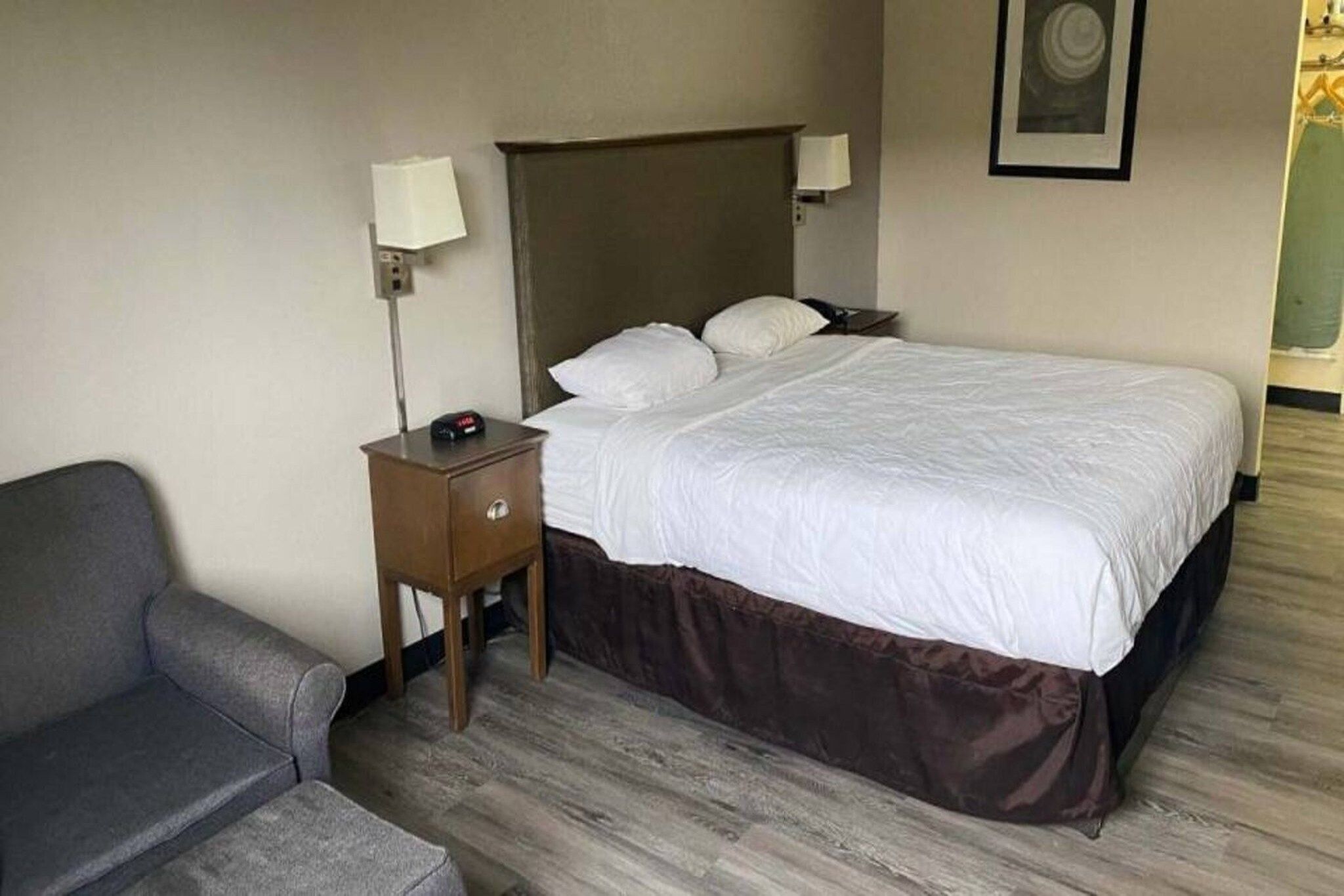 Une chambre d’hôtel avec un lit, une table de chevet, une lampe, une chaise et un tableau accroché au mur.