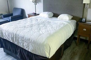 A hotel room with a large bed, a bedside table with a lamp, a chair, and a nightstand with a phone. - 1 King Bed | Smoking (Rochester NY)