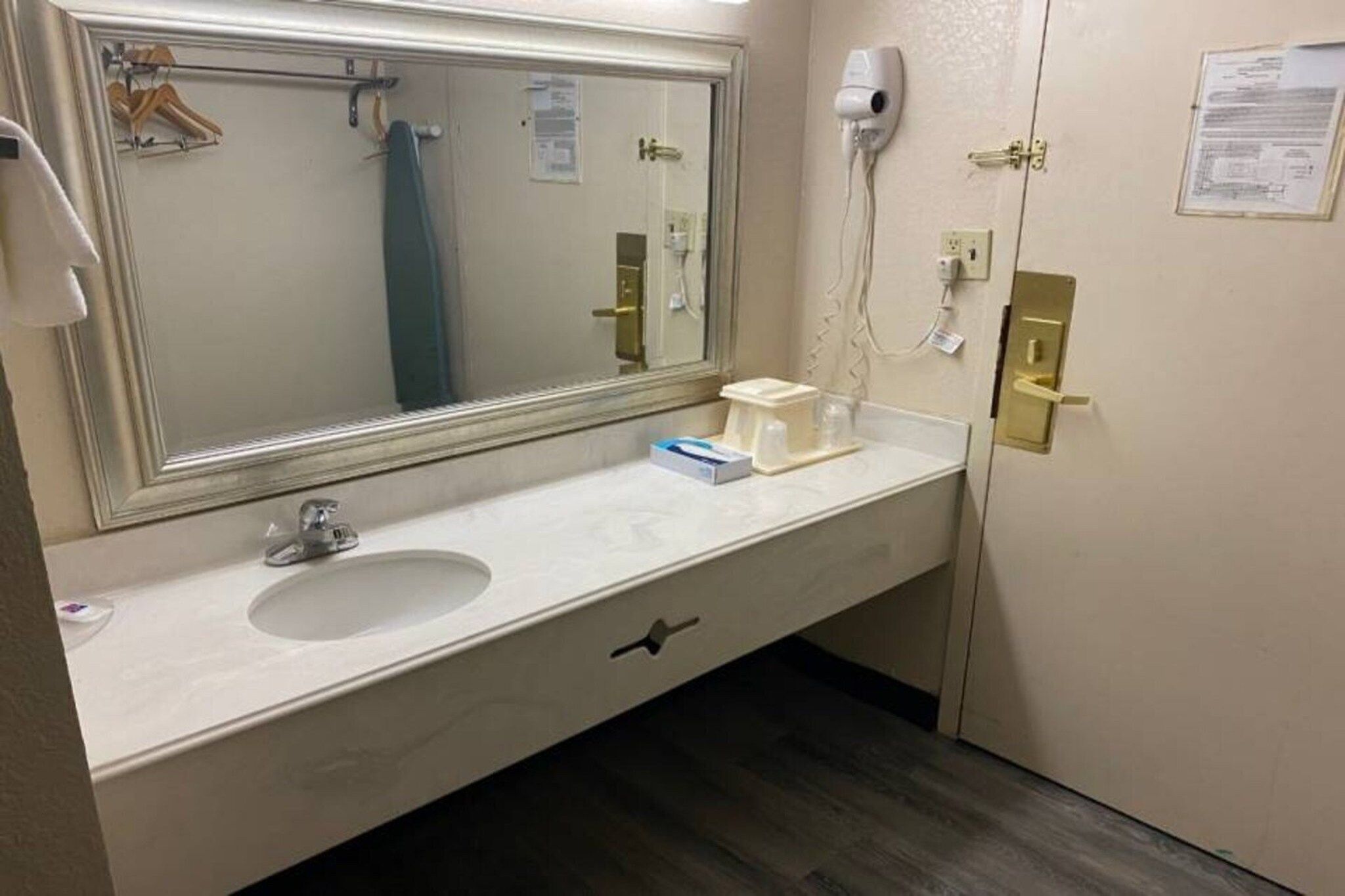 Een hotelbadkamer met een grote spiegel, een wastafel, een handdoekrek, een haardroger en een vel papier aan de muur.