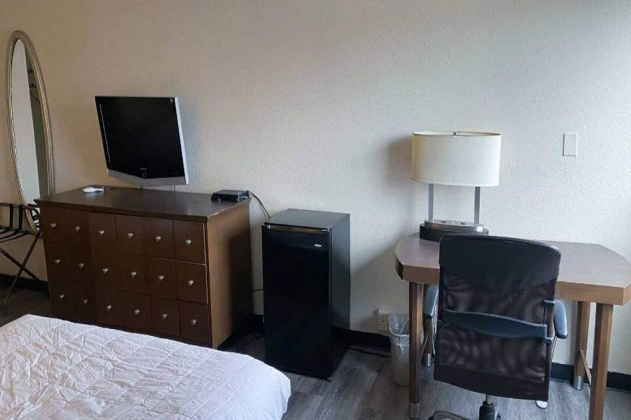 A hotel room with a bed, a dresser, a desk with a lamp, a television, and a refrigerator.
