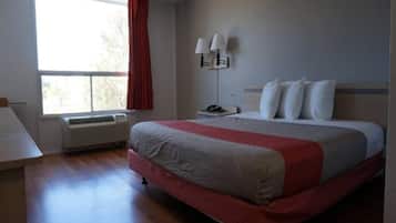 A hotel room with a large bed, a desk, a chair, a window with curtains, and a wall-mounted lamp.