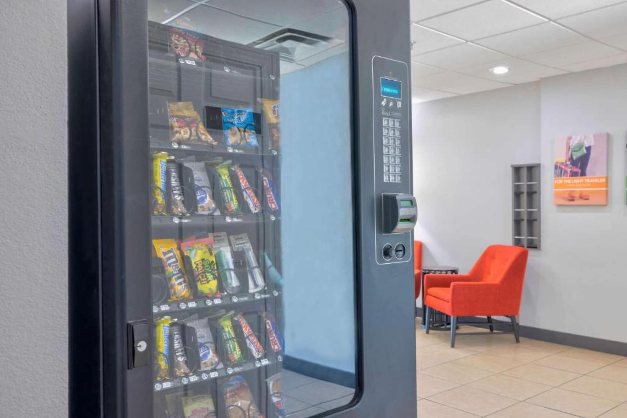 Máquina expendedora con aperitivos, un terminal de pago y una zona de estar con sillas naranjas.