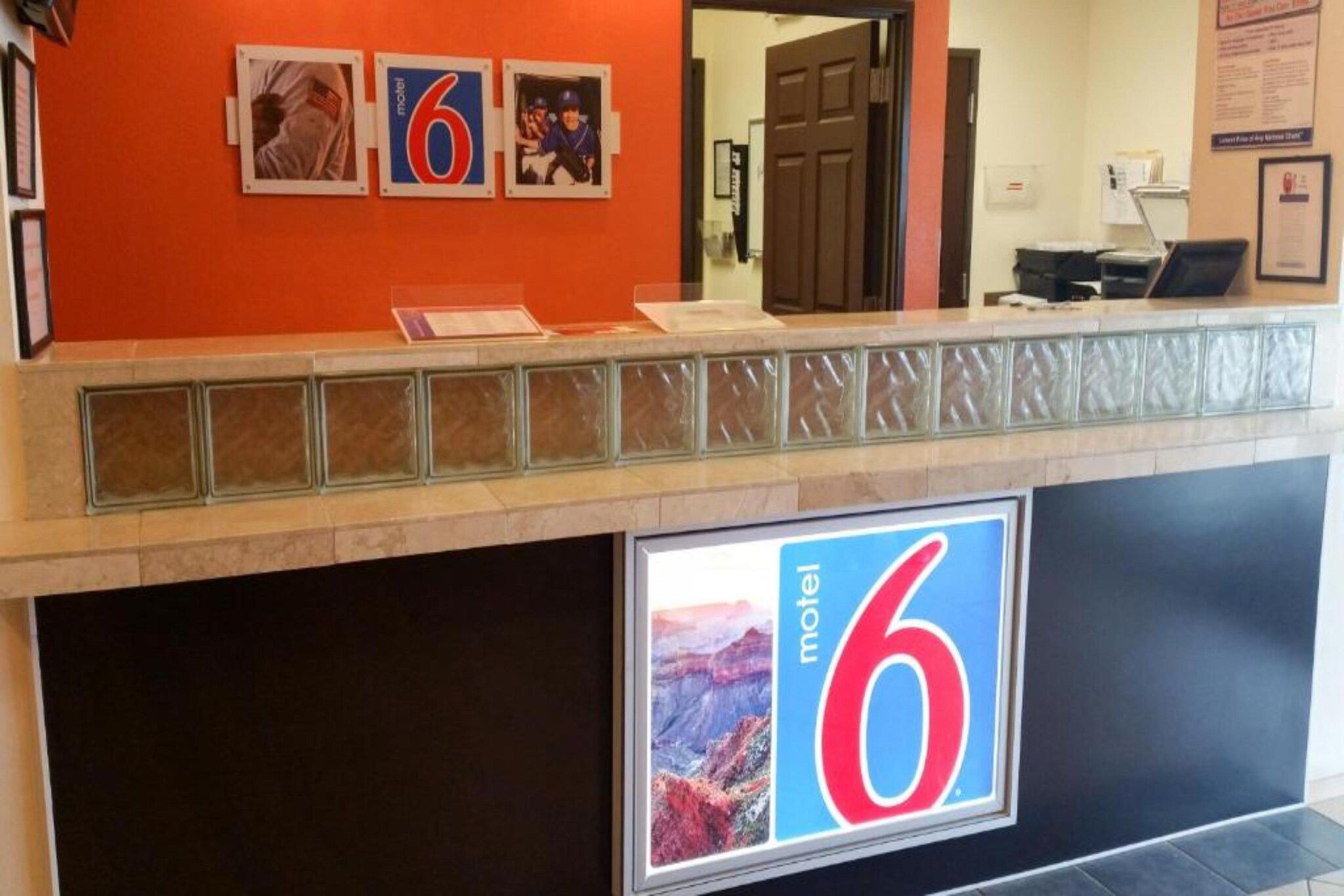 Mostrador de recepción de un motel con un cartel grande con el número '6', una mampara de vidrio y fotografías enmarcadas en la pared.