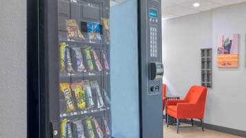 Ein Getränkeautomat mit Snacks und einem Bezahlsystem in einem Raum mit einem Stuhl und einem Wandplakat.
