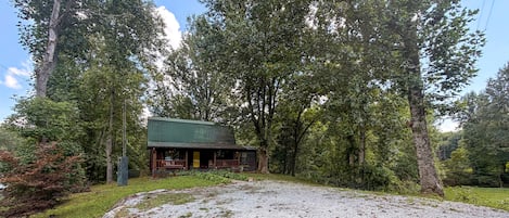A rustic cabin with a gravel driveway, surrounded by dense trees.