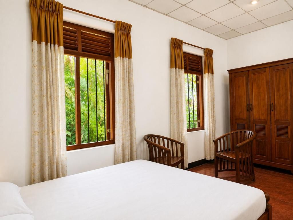 A hotel room with a bed, two chairs, a wardrobe, and windows with curtains.