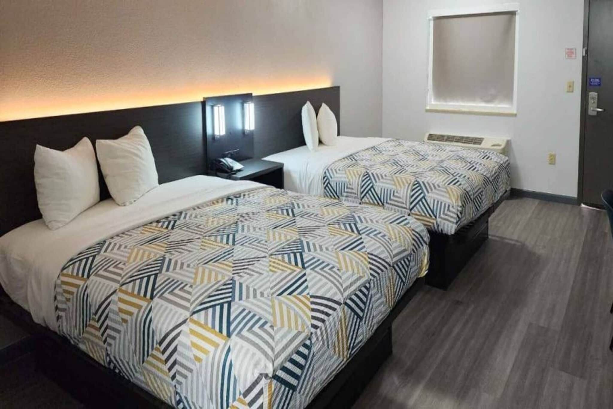 A hotel room with two beds, each with a patterned bedspread, a bedside table with a lamp, and a wall-mounted light fixture.
