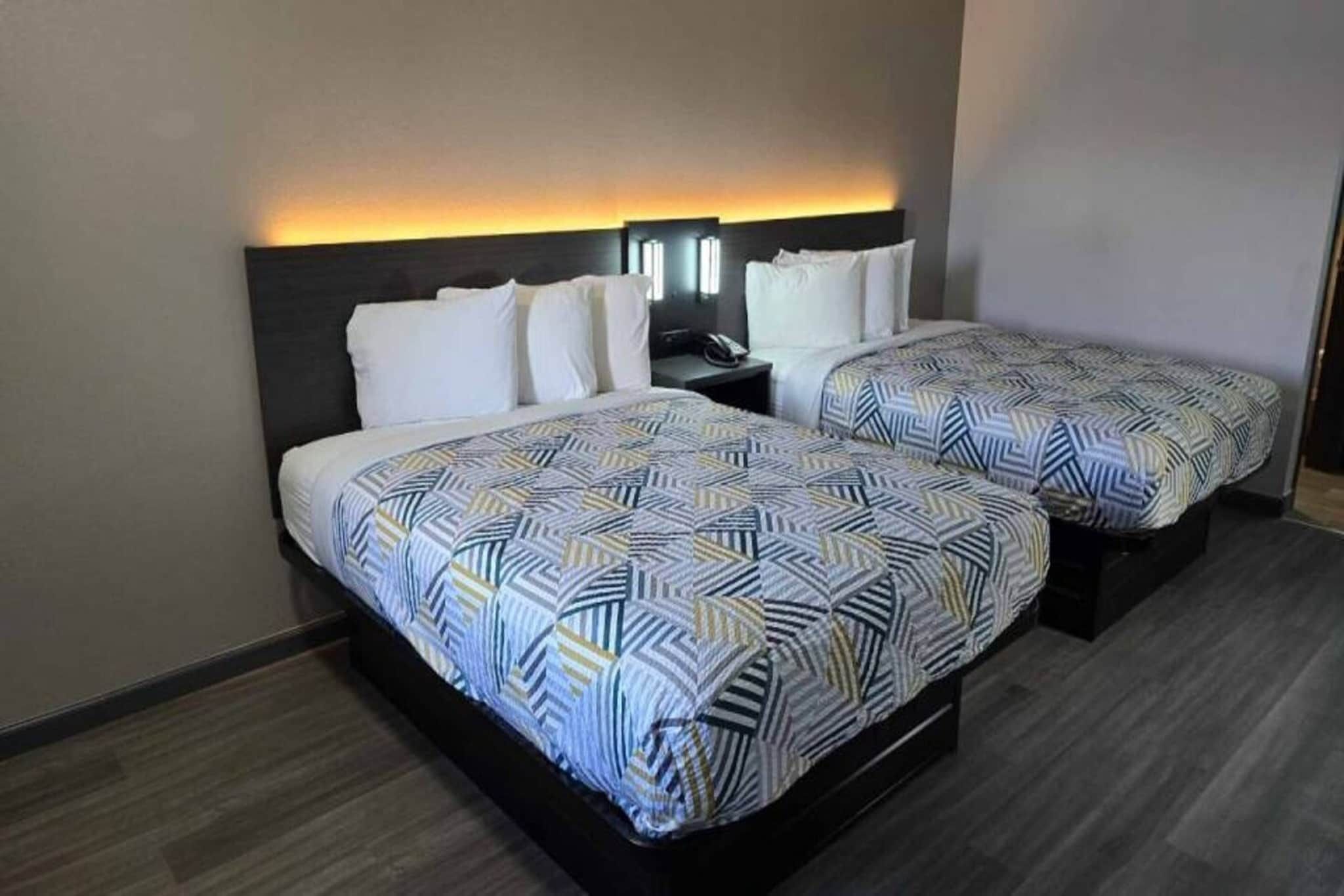 A hotel room with two beds, each with a patterned bedspread, a nightstand with a phone, and a wall-mounted light fixture.