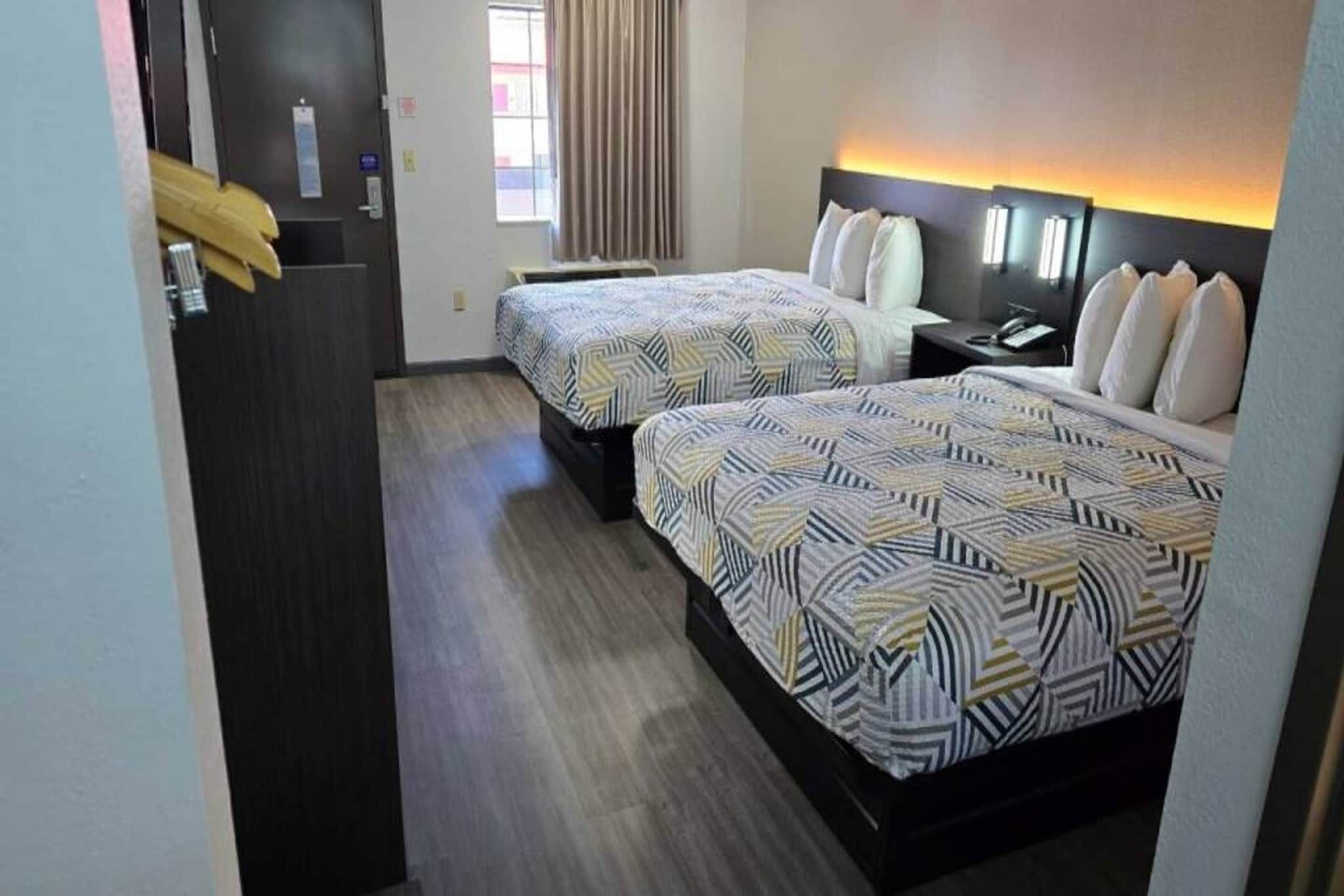 A hotel room with two beds, a wooden floor, a window with curtains, and a nightstand with a lamp.