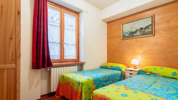 Een kamer met twee bedden, een raam met rode gordijnen, een radiator en een schilderij aan de muur.