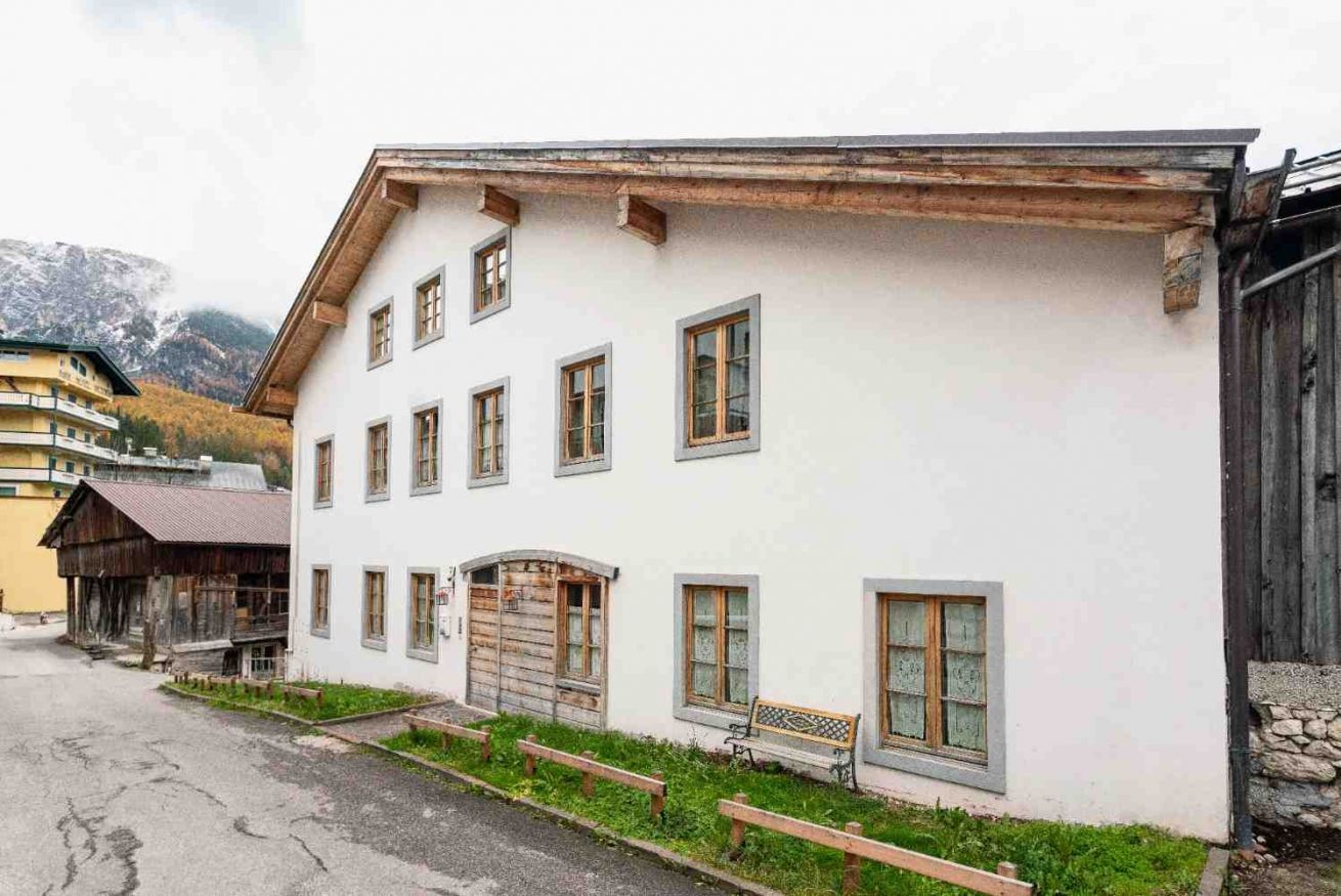 A white building with multiple windows and a wooden door, surrounded by a street and a mountain backdrop.