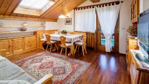 A cozy room with a wooden ceiling, a dining table with chairs, a TV, and a large window with curtains. - Apartment for 5 with Panoramic Terrace Near Cortina Centre (Cortina d'Ampezzo)