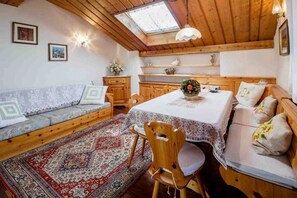 Une pièce chaleureuse avec un plafond en bois, une table à manger recouverte d’une nappe en dentelle, un canapé agrémenté de coussins à motifs et un tapis traditionnel.