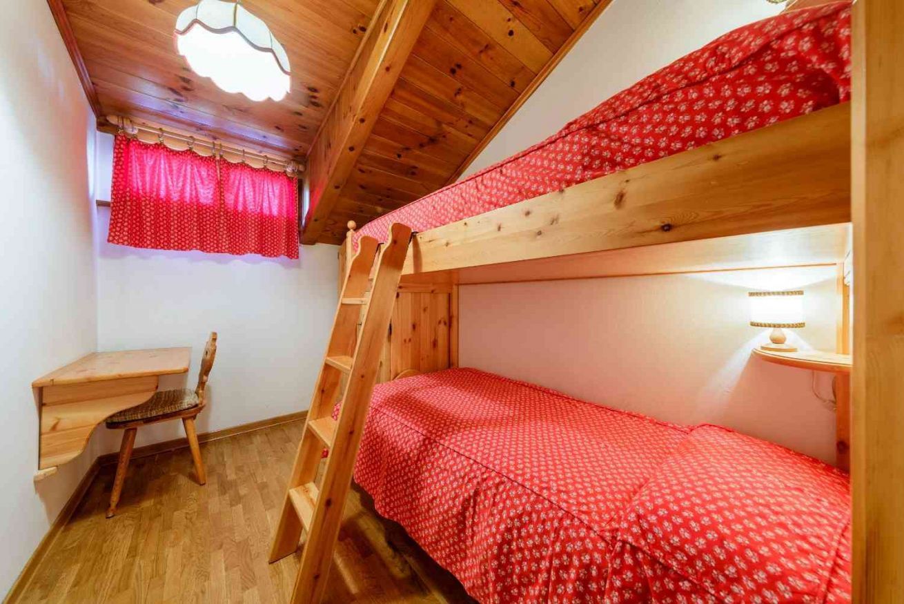 A bunk bed with a red patterned bedspread, a wooden desk with a chair, and a ladder leading to the upper bunk.