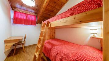 A bunk bed with a red patterned bedspread, a wooden desk with a chair, and a ladder leading to the upper bunk.