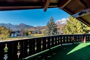 Un balcón con vistas a un paisaje montañoso, baranda de madera con detalles decorativos y un cielo azul despejado.