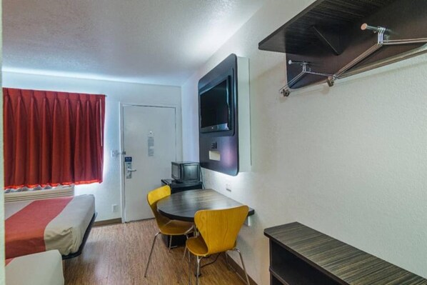 A compact room with a bed, a dining table with chairs, a television mounted on the wall, and a small cabinet.