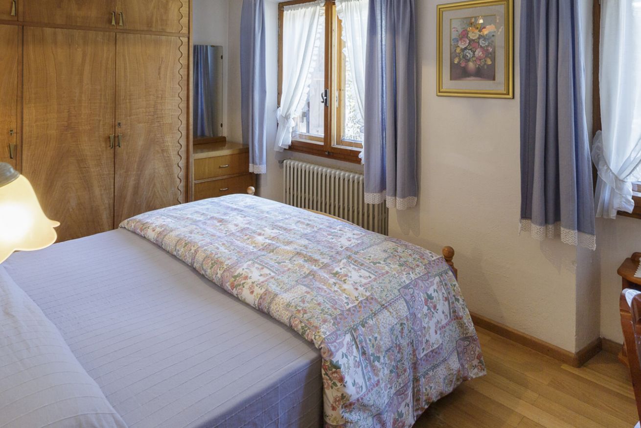 Un dormitorio con cama, armario de madera, ventana con cortinas, radiador y un cuadro en la pared.