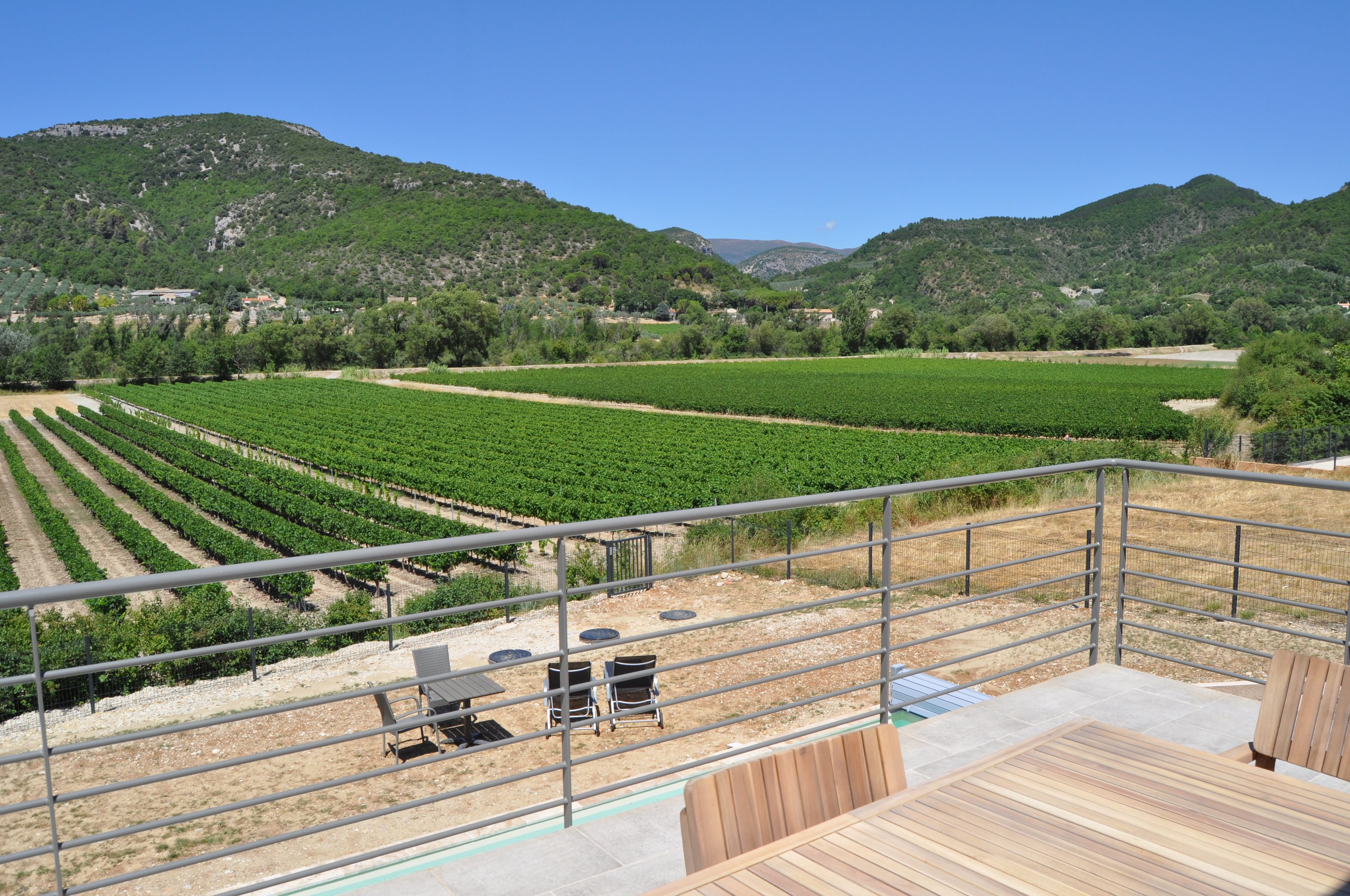 Un vignoble avec des rangées de vignes, une terrasse avec des chaises et des tables, et un paysage montagneux en arrière-plan.