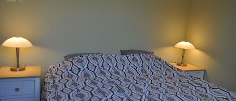 A bedroom with a patterned bedspread, two bedside lamps, a nightstand, and a wall-mounted light switch.