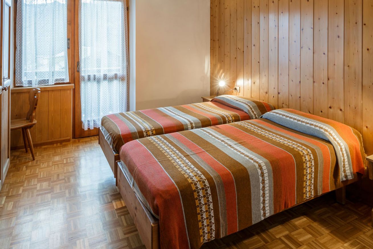 A room with two beds, wooden flooring, a window with sheer curtains, and a wall-mounted light.