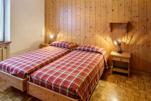 A room with two wooden beds, a nightstand with a lamp, and a wooden shelf.