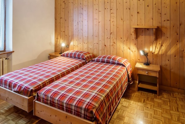 A room with two wooden beds, a nightstand with a lamp, and a wooden shelf.
