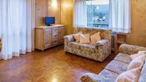 Una sala de estar con piso de madera, un sofá floral, un televisor sobre un mueble de madera y una ventana con vistas.