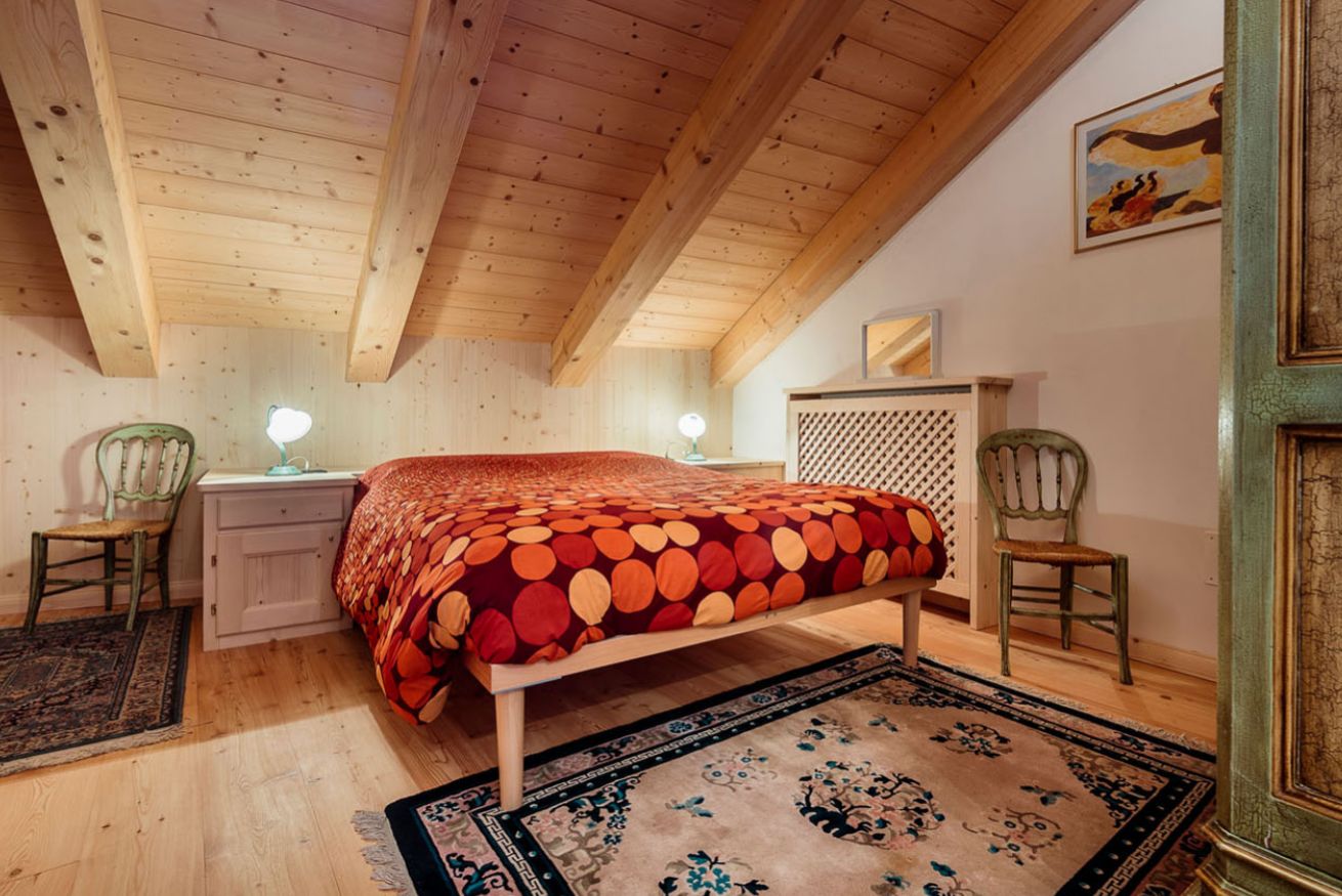 A bedroom with a wooden ceiling, a bed with a patterned bedspread, two bedside tables with lamps, and a decorative chair.