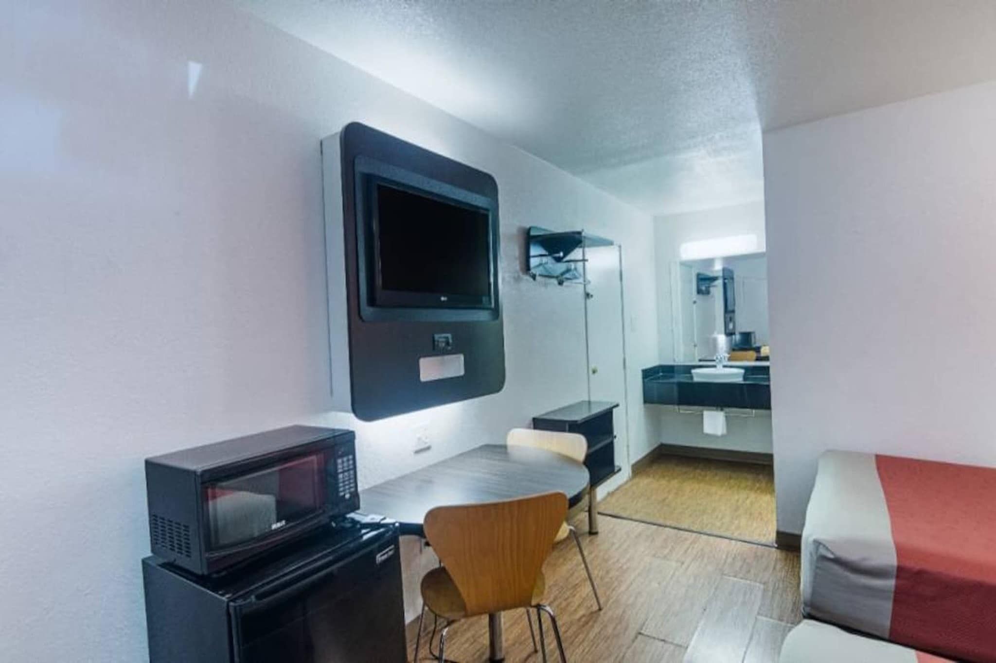 A compact room with a microwave, a small table, a chair, a sofa, a TV mounted on the wall, and a mirror above the sink.