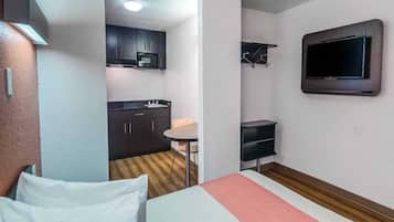 Habitación de hotel compacta con kitchenette, cama con ropa blanca, una mesita y televisor montado en la pared.