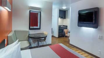 Habitación de hotel con cama, una mesita, una silla, televisión y una kitchenette al fondo.