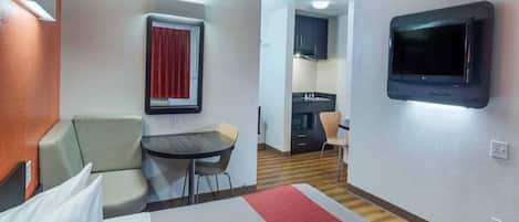 A hotel room with a bed, a small table, a chair, a TV, and a kitchenette in the background.