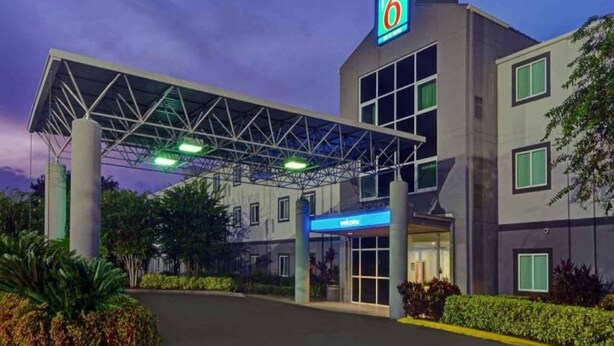 A hotel with a covered entrance, a prominent '6' logo, and a blue welcome sign. - 2 Full Beds | Premium, Mini Fridge (Orlando FL)