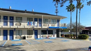 Um motel de dois andares com marquise azul, um letreiro com 'Ofertas Semanais' e uma placa de 'Bem-vindo' acima da entrada.
