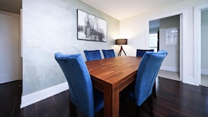 Un coin repas avec une table en bois et des chaises bleues, une œuvre d ’art représentant un paysage urbain accrochée au mur, et une porte donnant sur une autre pièce.